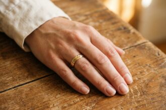 Alliance en or jaune portée sur une main de femme posée sur une table en bois, gros plan naturel pour illustrer le choix d'une alliance de mariage classique